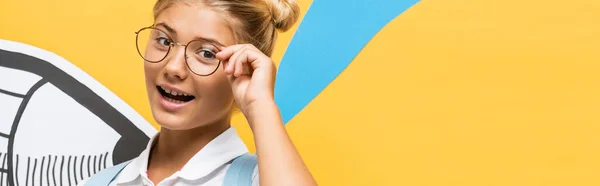 全景概念快乐的女学生戴着眼镜 望着近纸的相机 用蓝色和黄色裁剪铅笔 — 图库照片