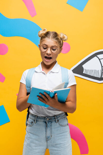 excited schoolgirl in eyeglasses reading book on yellow background with paper pencil and multicolored elements