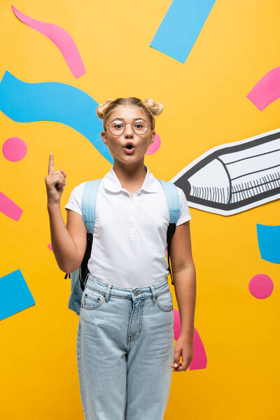 amazed schoolgirl in eyeglasses showing idea sign near paper pencil and abstract elements on yellow