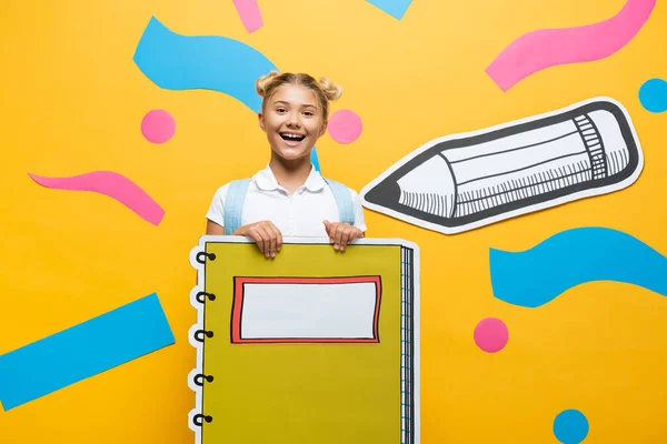 Joyful Schoolgirl Holding Notebook Maquette Looking Camera Yellow Background Paper — Stock Photo, Image