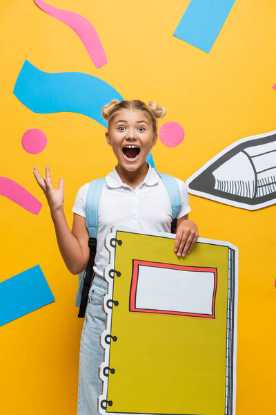 excited schoolgirl with notebook maquette screaming and gesturing on yellow background with decorative elements and paper cut pencil