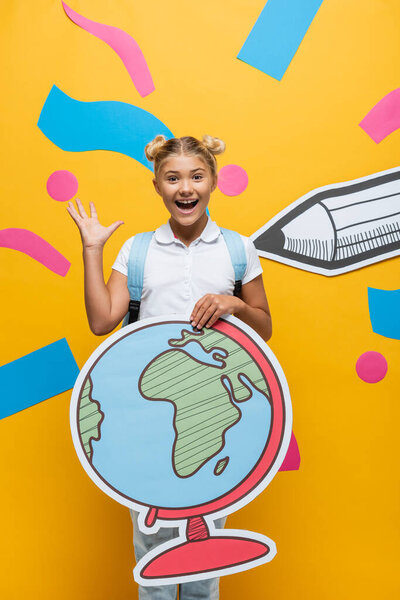 joyful schoolgirl waving hand while holding globe maquette on yellow background with paper cut pencil and colorful elements 
