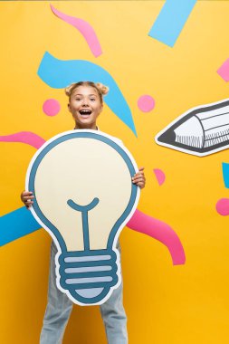 Excited schoolgirl holding paper light bulb beside paper elements on yellow background clipart