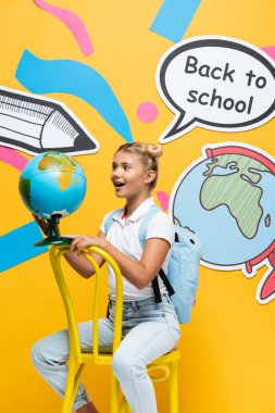 Heyecanlı okul çocuğu sarı arka planda kağıdın yanındaki küreye bakıyor.