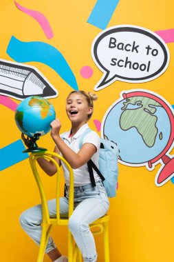 Excited schoolgirl holding globe near speech bubble with back to school lettering and paper artwork on yellow background clipart