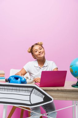 Çocuk pembe arka planda dizüstü bilgisayar, küre ve kağıt sanatının yanında oturuyor.