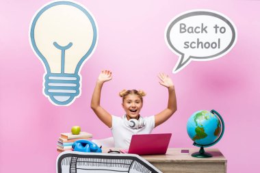 Kulaklıklı okul çocuğu pembe arka planda cihazlar, dünya ve kağıt işlerinin yanında oturuyor.