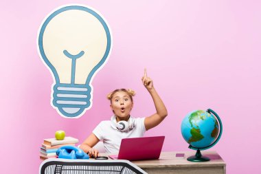 Heyecanlı okul çocuğu, pembe arka planda cihazların ve kağıt sanatının yanında fikir yürütüyor.