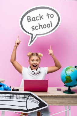 Heyecanlı çocuk okula geri dönen konuşma baloncuğunu işaret ediyor dizüstü bilgisayar, akıllı telefon ve pembe arka plandaki küreyi gösteriyor.