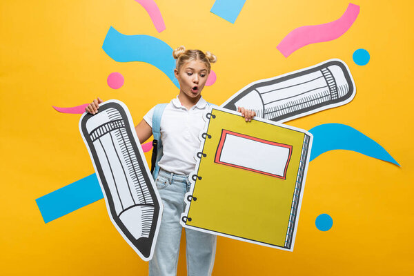Shocked schoolchild holding paper notebook and pencil on yellow background