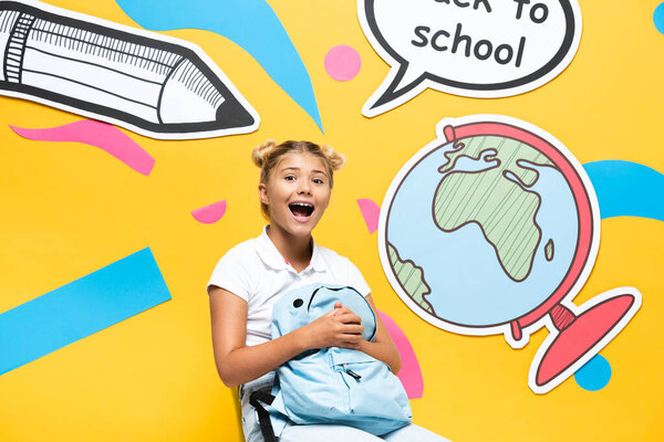 Excited schoolgirl holding backpack near speech bubble and paper craft  on yellow background