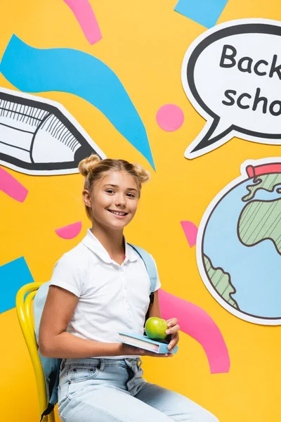 Pupil holding book and apple on chair near paper speech bubble on yellow background