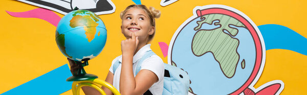 Панорамный снимок мечтательного школьника с земным шаром, сидящего рядом с бумажными рисунками на желтом фоне