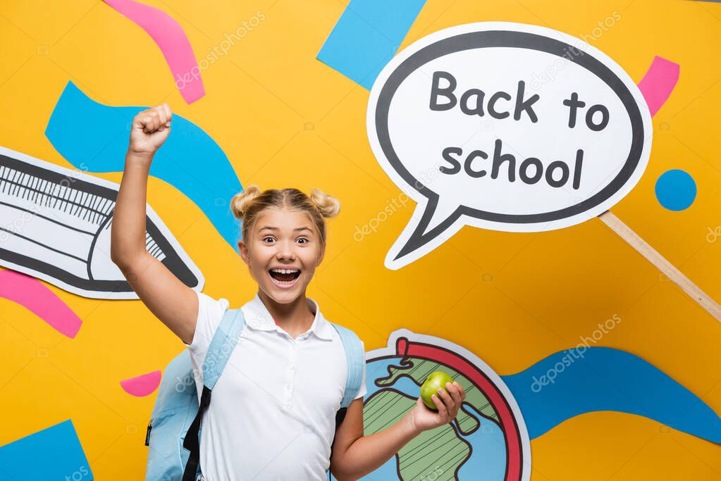 Excited schoolgirl with apple showing yeah gesture near speech bubble with back to school lettering and paper art on yellow background