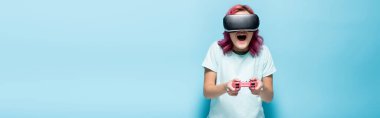KYIV, UKRAINE - 29 Temmuz 2020: VR kulaklıklı pembe saçlı heyecanlı genç kadın mavi arka planda joystick ile video oyunu oynuyor, panoramik çekim