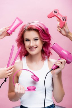 KYIV, UKRAINE - 29 Temmuz 2020: Düz saç, kulaklık, kozmetik krem, joystick ve saç kurutma makinesi yanında renkli saçlı genç bir kadın pembe ellerde izole edilmiş.