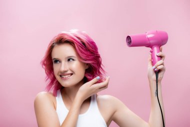 Renkli saçlı, saç kurutma makinesi tutan ve pembe üzerinde sırıtan genç bir kadın.