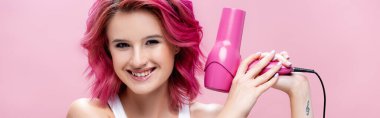 Pembe panoramik bir karede saç kurutma makinesi tutan, renkli saçlı genç bir kadın.