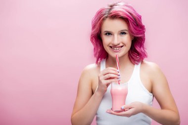 Renkli saçlı, çilekli milkshake 'i tutan ve pembe üzerinde saman içen genç bir kadın.