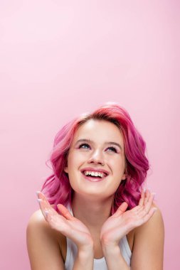 Renkli saçlı ve makyajlı genç bir kadın. Yüzüne yakın ellerle poz veriyor ve pembe bir şekilde gülümsüyor.