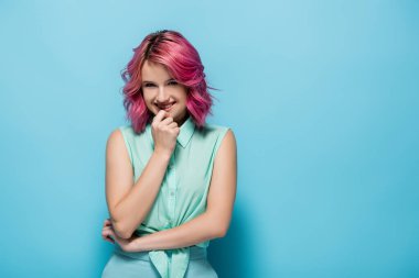 Pembe saçlı, gülümseyen ve mavi arka planda flört eden genç bir kadın.