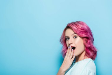 Şok olmuş, mavi arka planda pembe saçlı genç bir kadın.