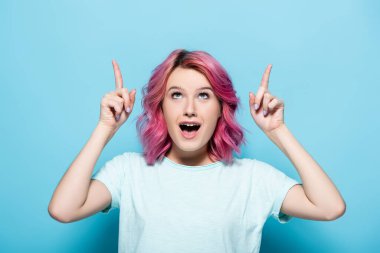 Şok olmuş, mavi arka planda pembe saçlı genç bir kadın.