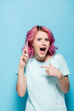 Şaşırmış, pembe saçlı genç bir kadın mavi arka planda akıllı telefonu işaret ediyor.