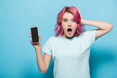 shocked young woman with pink hair holding smartphone with blank screen on blue background clipart