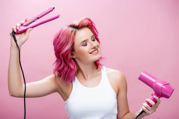 young woman with colorful hair and closed eyes holding straightener and hairdryer isolated on pink