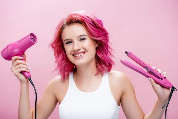 young woman with colorful hair holding straightener and hairdryer isolated on pink