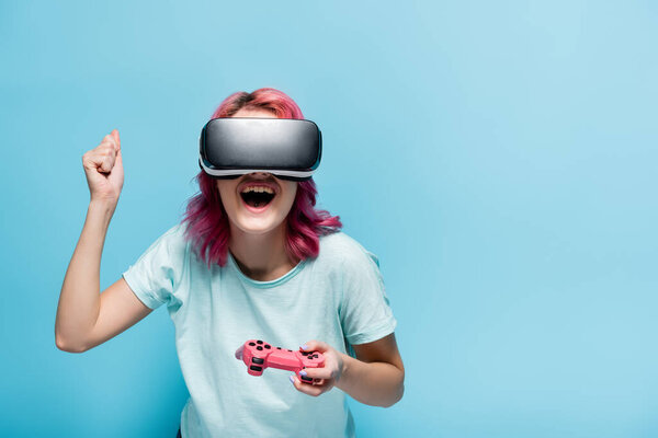 KYIV, UKRAINE - JULY 29, 2020: excited young woman with pink hair in vr headset with joystick on blue background