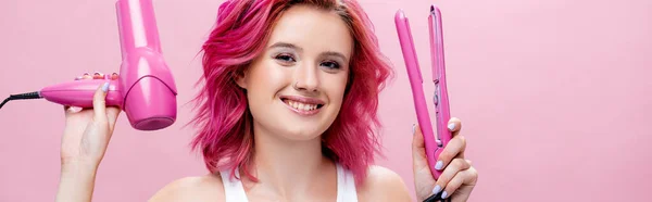 young woman with colorful hair holding straightener and hairdryer isolated on pink, panoramic shot