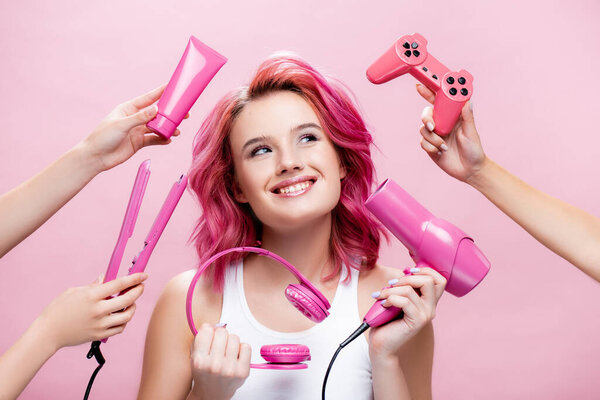 КИЕВ, Украина - 29 июля 2020 года: молодая женщина с красочными волосами возле выпрямителя, наушники, косметический крем, джойстик и фен в руках изолированы на розовый