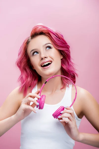 dreamy young woman with colorful hair holding headphones and looking up isolated on pink