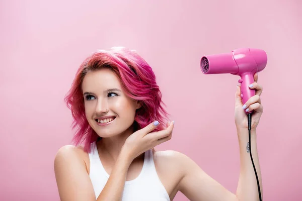 young woman with colorful hair holding hairdryer and smiling isolated on pink