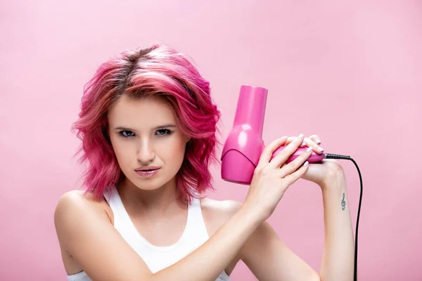 serious young woman with colorful hair holding hairdryer isolated on pink