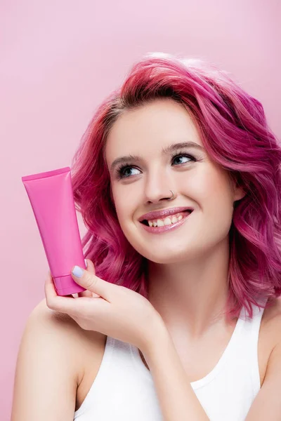 young woman with colorful hair holding tube of cosmetic cream isolated on pink