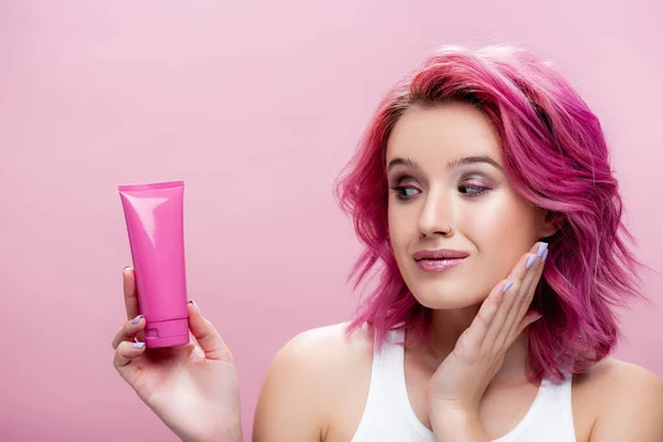 young woman with colorful hair looking at tube of cosmetic cream isolated on pink