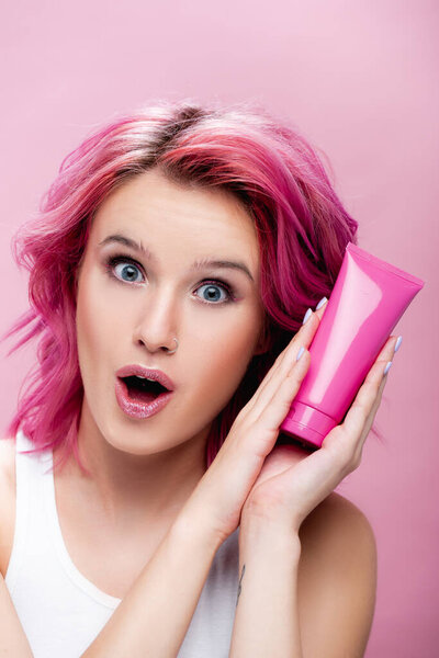 surprised young woman with colorful hair holding tube of cosmetic cream isolated on pink