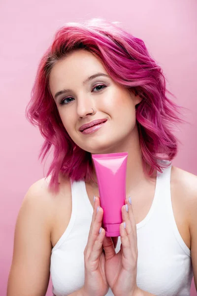 young woman with colorful hair holding tube of cosmetic cream isolated on pink