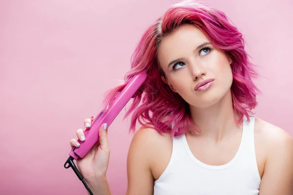 dreamy young woman with colorful hair using straightener isolated on pink