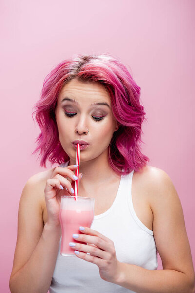 young woman with colorful hair drinking strawberry milkshake from straw isolated on pink