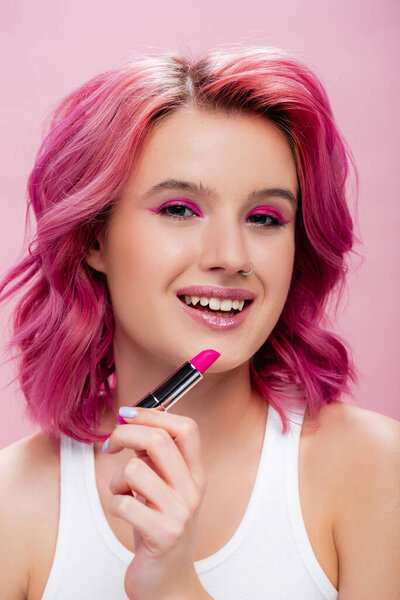 young woman with colorful hair holding lipstick and smiling isolated on pink