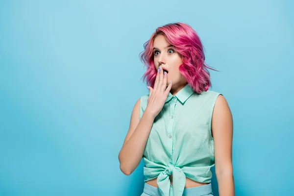 shocked young woman with pink hair covering mouth on blue background