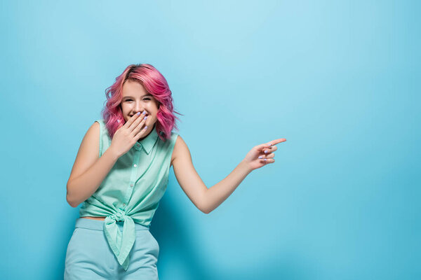 young woman with pink hair pointing aside and laughing on blue background
