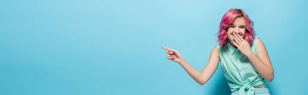 young woman with pink hair pointing aside and laughing on blue background, panoramic shot