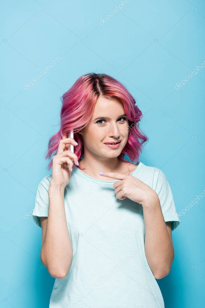 young woman with pink hair pointing at smartphone on blue background
