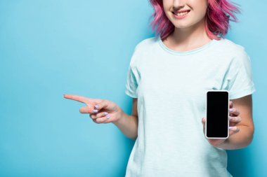 Pembe saçlı, elinde akıllı bir telefonla mavi arkaplanı işaret eden genç bir kadın görüntüsü.