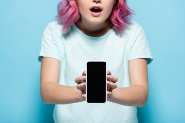 Şok olmuş, pembe saçlı, elinde akıllı bir telefonla mavi arka planda boş ekranlı bir kadın görüntüsü.
